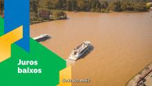 Imagem aérea de dois barcos turisticos navegando em um trecho de rio. à esqueda, sobre um fundo em grafismo verde aparede o texto juros baixos e embaixo uma legenda do audio escrito "cada vez mais"