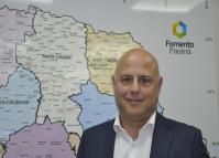 Claudio Pacheco é um homem branco, calvo, usa camisa branca e paletó azul marinho, tem ao fundo um mapara o Paraná com uma logomarca da Fomento Paraná