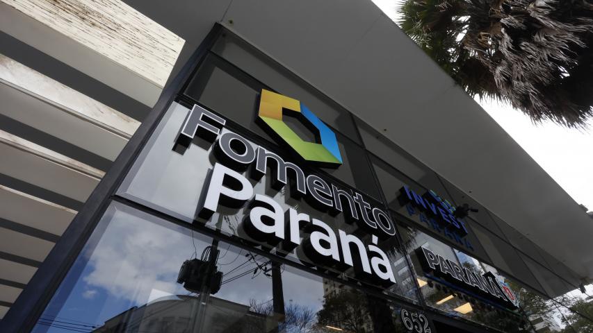Parede envidraçada da fachada do edifício sede da Fomento Paraná, mostra logomarca da instituição em primeiro plano, formada por um hexágono nas cores azul, amarelo e verde.