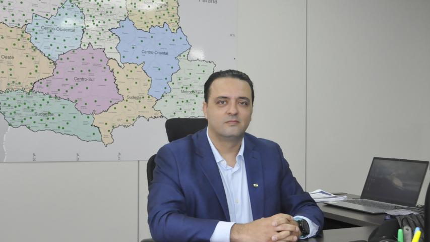 Gustavo Emanuel Cejas é um homem branco, usa terno azul escuro e camisa azul clara. Está sentado em uma mesa de escritório. Há um mapa do Paraná na parede atrás dele e um lap top na mesa ao lado.